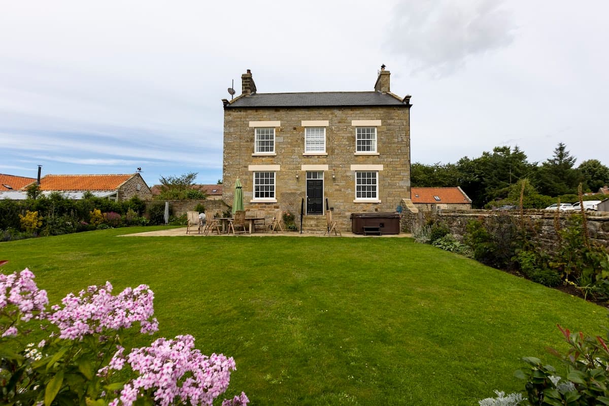 Stone cottage with a manicured lawn, surrounded by flowers and trees, featuring a patio and hot tub.