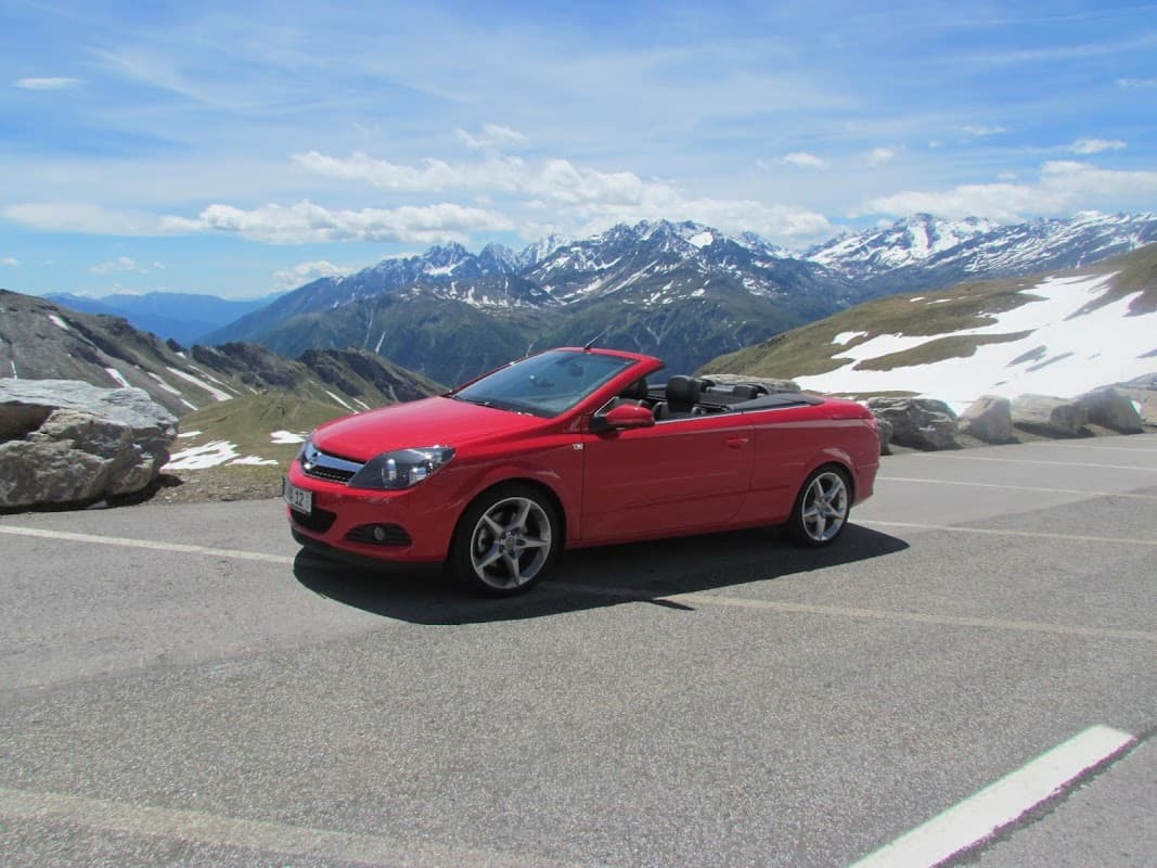 Red convertible car parked on a mountain road with snow-capped peaks and blue sky in the background.