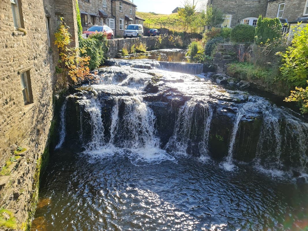 A serene waterfall cascades over rocks, surrounded by stone buildings and greenery in Hawes, Yorkshire.