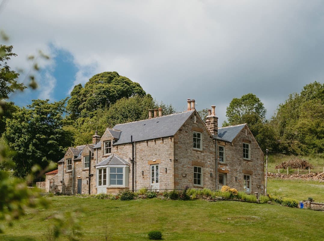 Stone building with multiple windows, surrounded by greenery and trees under a partly cloudy sky.