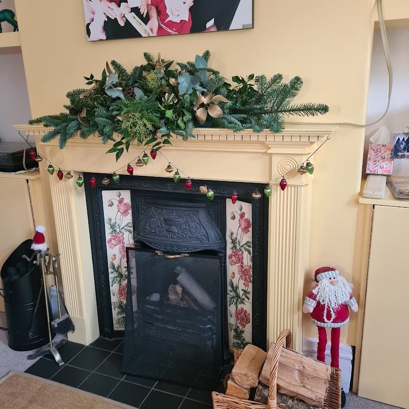 Festive mantel decorated with greenery, ornaments, and a Santa figure beside a black fireplace and wood basket.