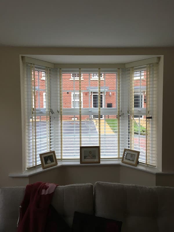 Bay window with white blinds, framed pictures on the sill, and a view of a brick building and garden outside.