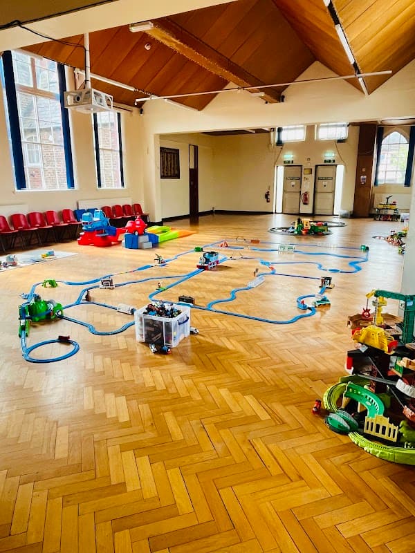 Bright hall with wooden flooring, toy train tracks, colorful playsets, and seating along the walls.