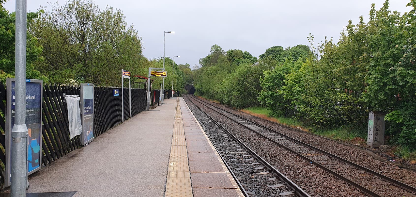 Car park beside railway tracks, trees lining the area, and platform with signage under overcast sky.