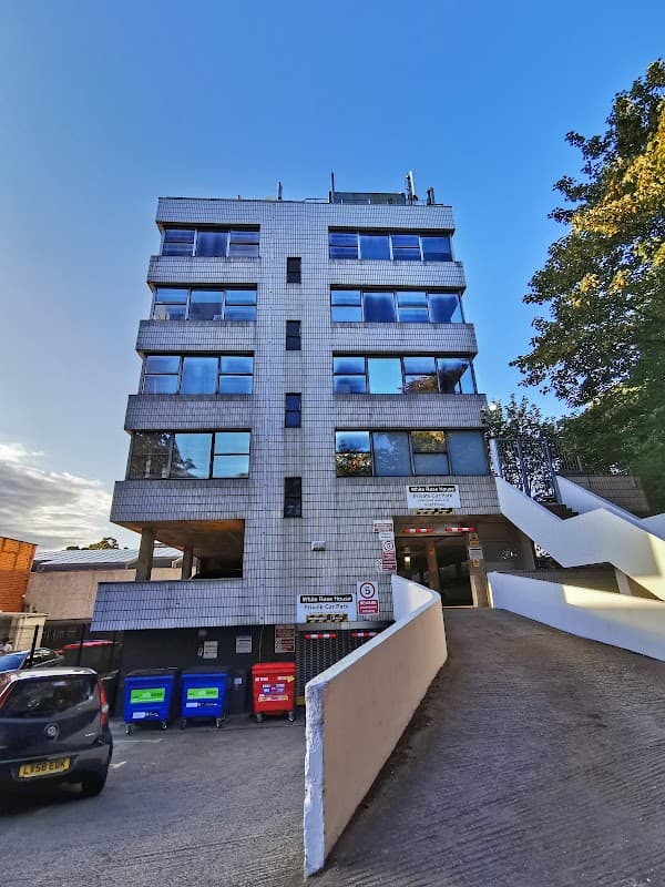 Modern building with large windows, a sloped driveway, and recycling bins in a private car park setting.