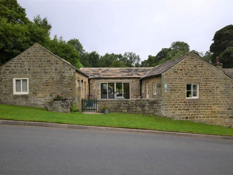 Charming stone cottage with a sloped lawn, green gate, and large windows, set in a rural North Yorkshire landscape.