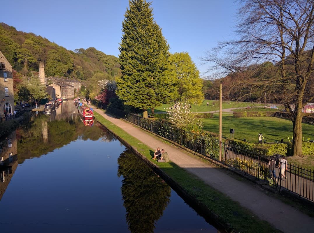 Scenic view of a canal reflecting trees, with people walking along the path and a park in the background in Hebden Bridge.