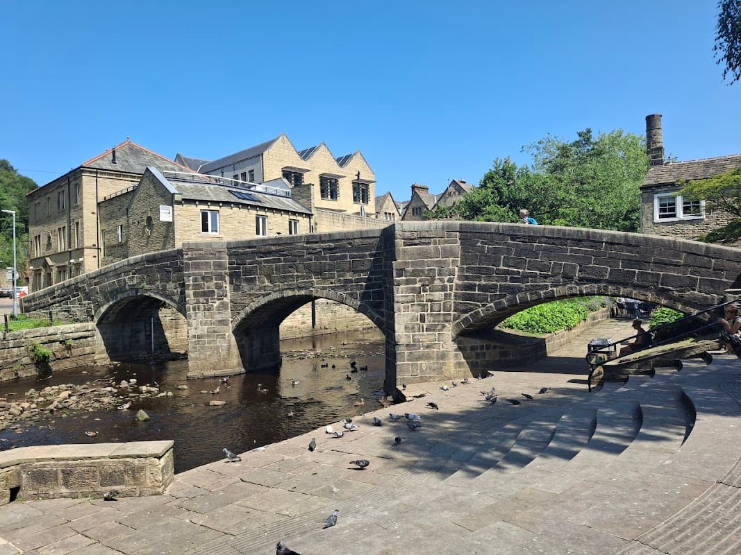 Stone packhorse bridge over a river, surrounded by historic buildings and greenery, with people and pigeons nearby.