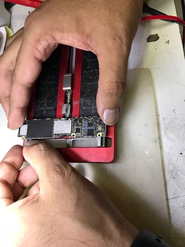 Hands holding a circuit board over a red repair tool, with tools and a work surface visible in the background.