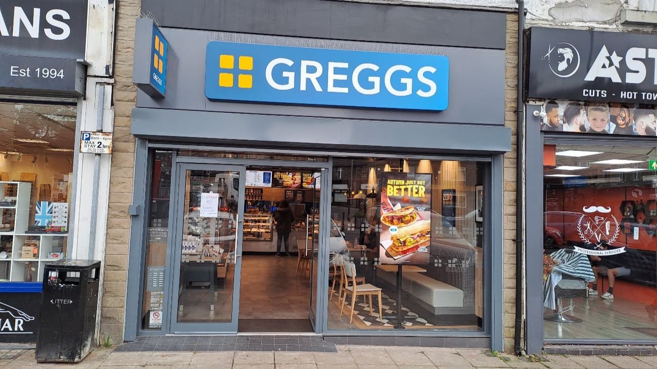 Brightly lit Greggs shop front with large signage, displaying food items and seating visible inside.