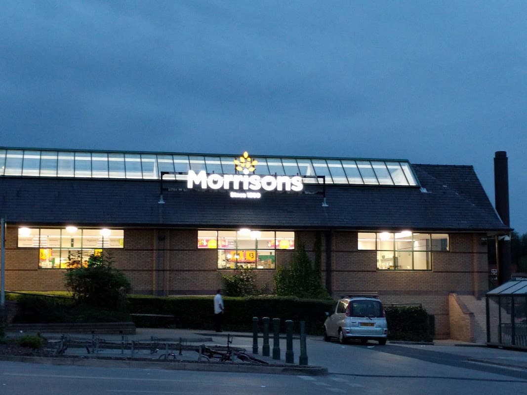 Morrisons store in Heckmondwike, Yorkshire, illuminated at dusk with a clear sky and parking area in front.