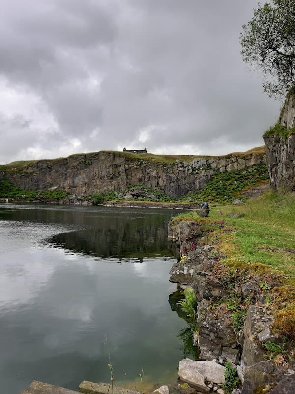 Serene waters reflect rugged cliffs under a cloudy sky, with a distant building atop the hill at Helwith Bridge.