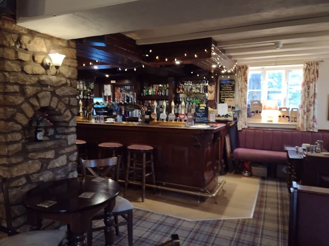 Cozy bar interior with wooden counter, stone wall, stools, and warm lighting, featuring shelves of drinks and a plaid carpet.