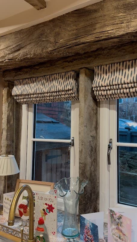 Wooden beams frame two windows adorned with patterned blinds, surrounded by decorative cards and a glass vase.