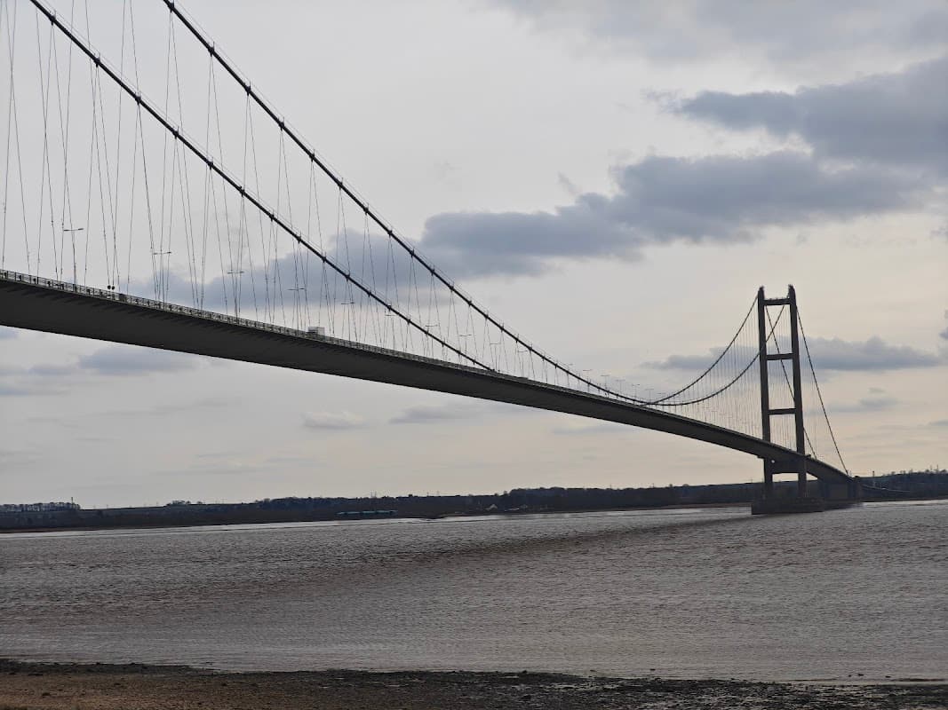 The Humber Bridge stretches across a cloudy sky, towering over the muddy riverbanks below.