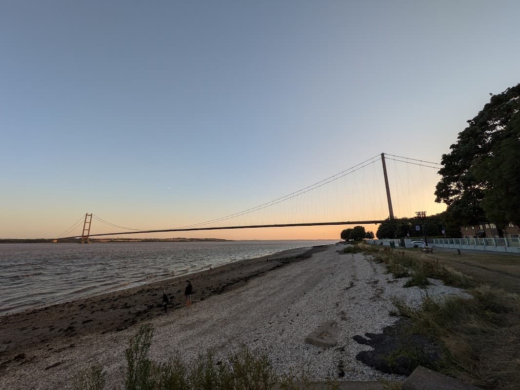 Foreshores East Car Park with a view of the Humber Bridge at sunset, sandy shore, and greenery nearby.