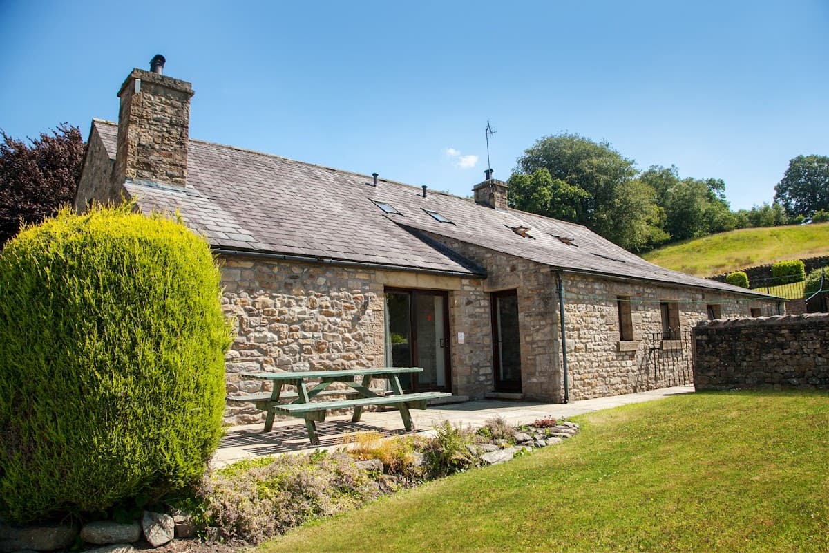 Stone building with a slate roof, picnic table, and lush green lawn surrounded by trees in a sunny landscape.
