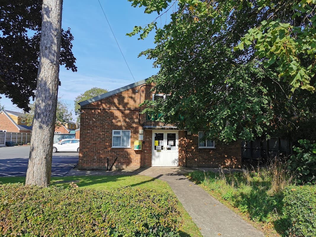 Brick building with a white door, surrounded by greenery and trees, set against a clear blue sky.