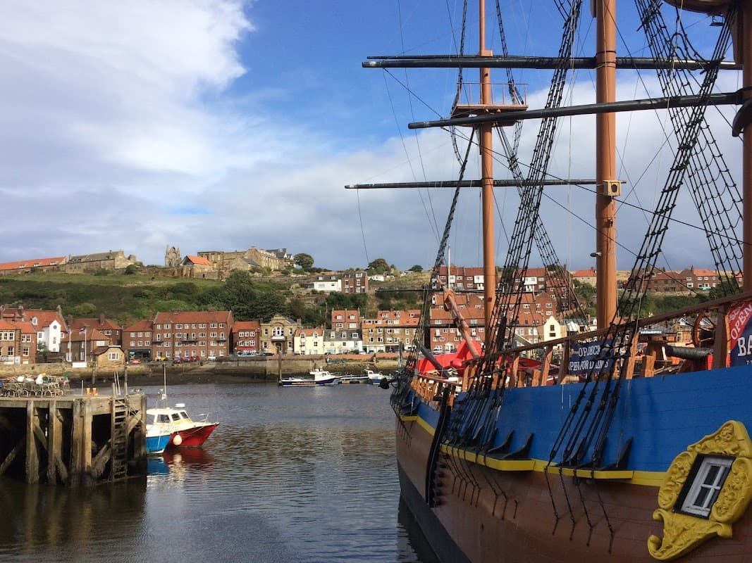 A dock with a wooden ship, colorful buildings on the hillside, and a blue sky with scattered clouds.