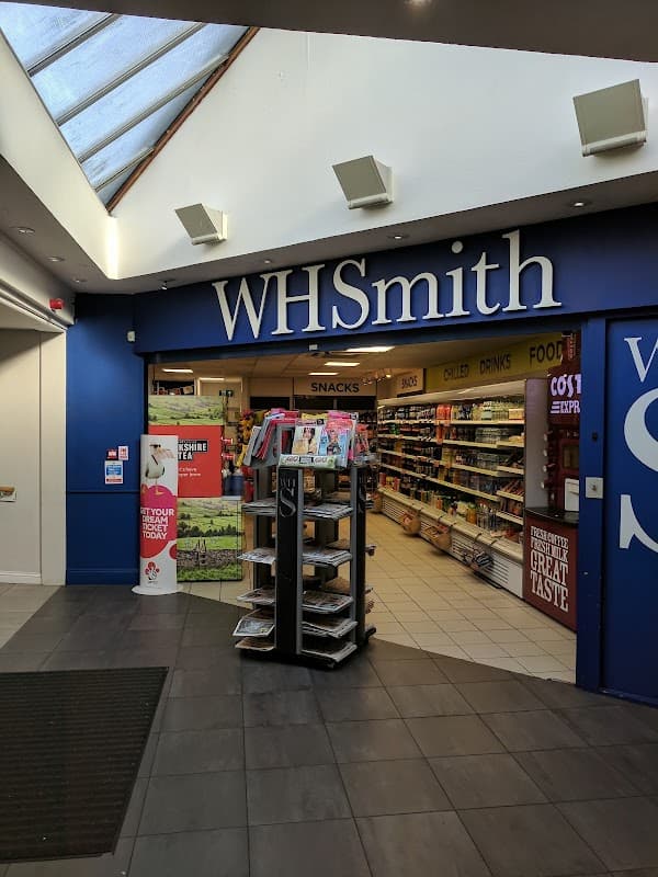 WHSmith storefront with blue signage, shelves stocked with snacks and drinks, and a display stand for magazines.
