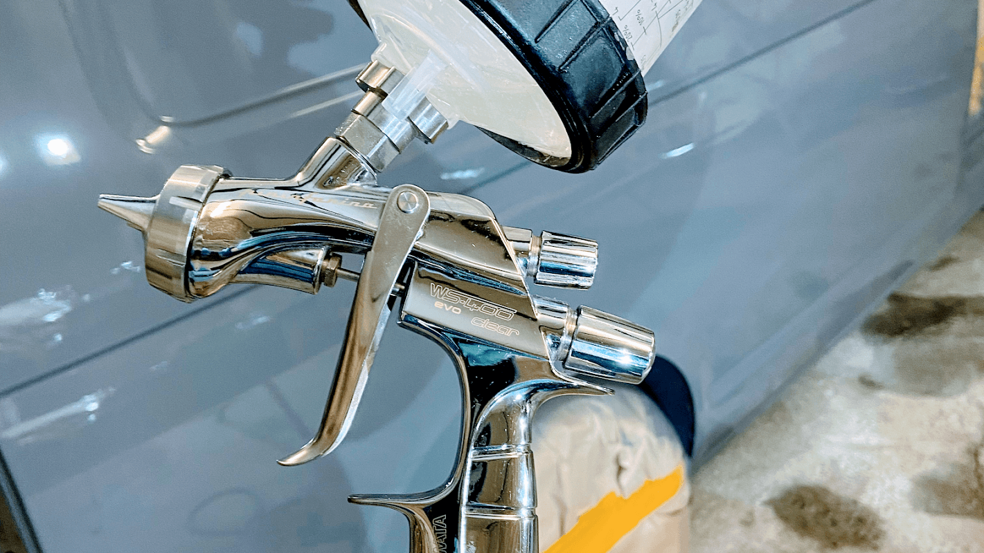 Close-up of a chrome spray gun with a paint cup, set against a blurred background of a workshop in Hinderwell, Yorkshire.