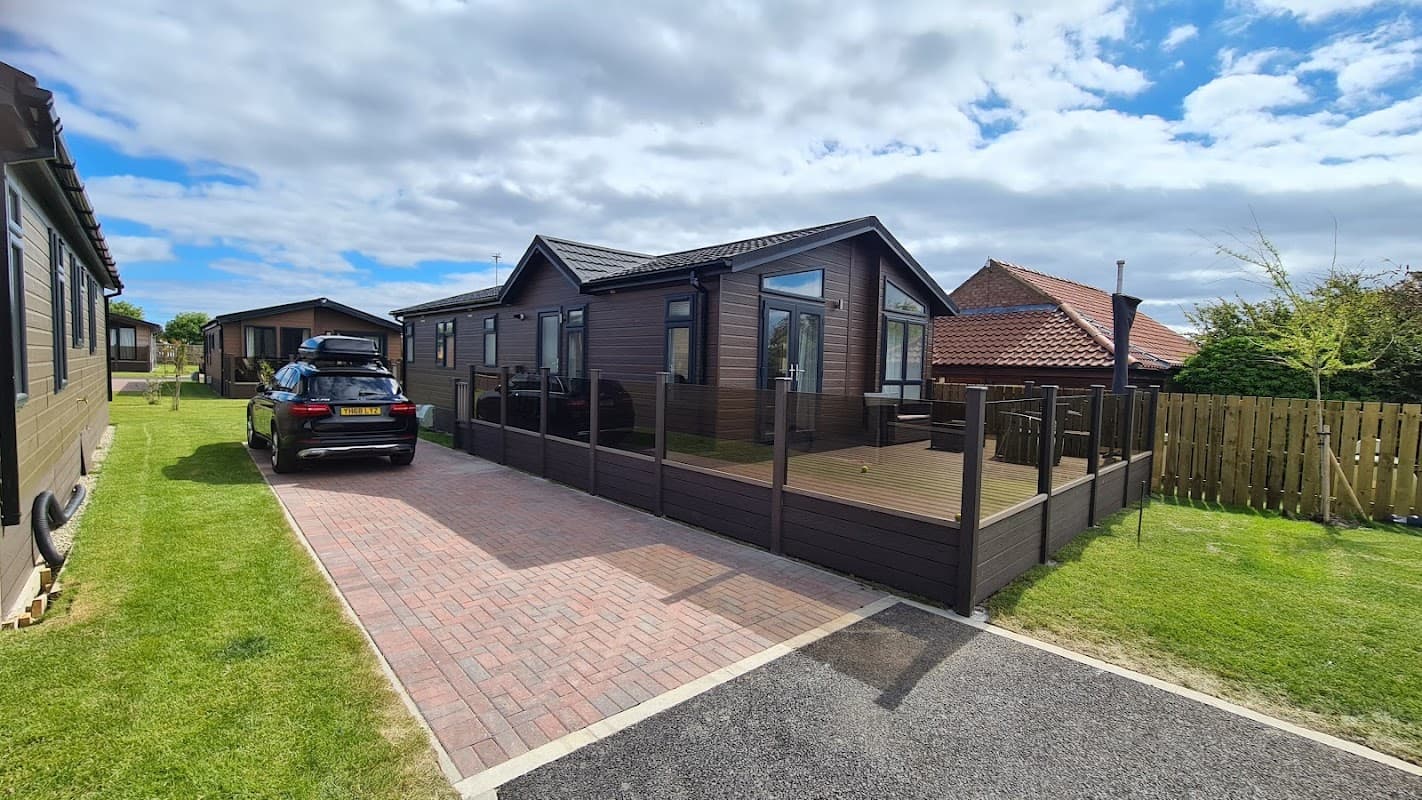 Modern holiday lodges with wooden exteriors, a paved driveway, and green lawns under a partly cloudy sky.