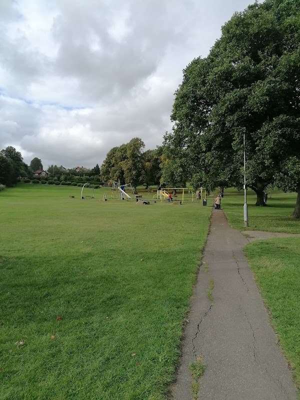 Lush green park with a pathway, playground equipment, and trees under a cloudy sky in Hipperholme, Yorkshire.