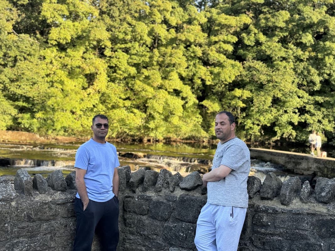 Two men stand by a stone wall near a river with lush green trees in the background, enjoying a sunny day.