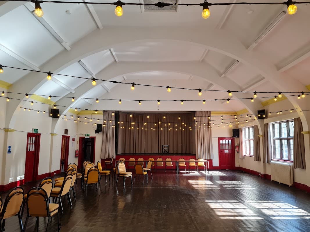 Brightly lit hall with string lights, rows of chairs, and a stage area, featuring large windows and wooden flooring.