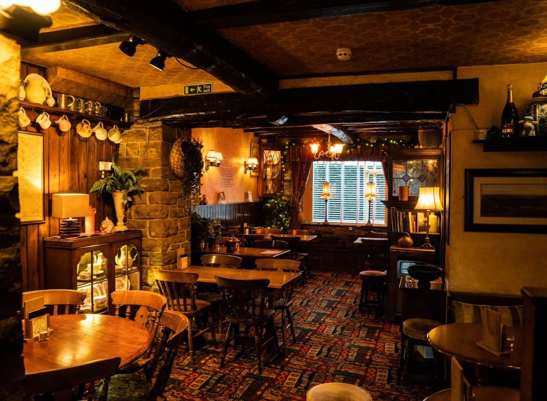 Cozy bar interior with wooden beams, tables, chairs, and warm lighting, featuring a stone fireplace and decorative shelves.