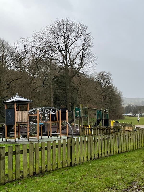 Playground equipment behind a wooden fence, with trees and a cloudy sky in the background at Sands Recreation Ground.