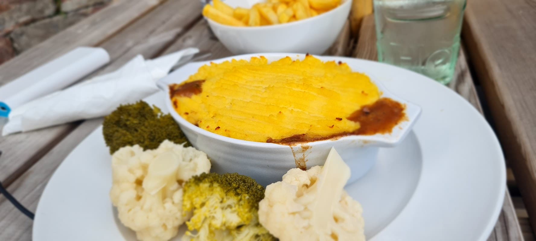 Shepherd's pie with a golden topping, served with cauliflower and broccoli, alongside a bowl of fries.
