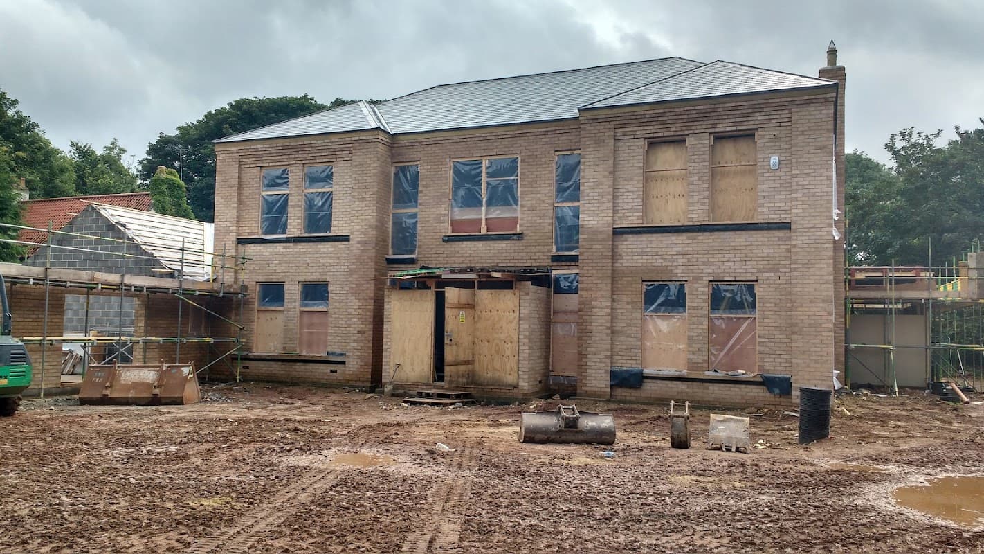 Two-story brick building under construction with boarded windows, surrounded by muddy ground and construction materials.