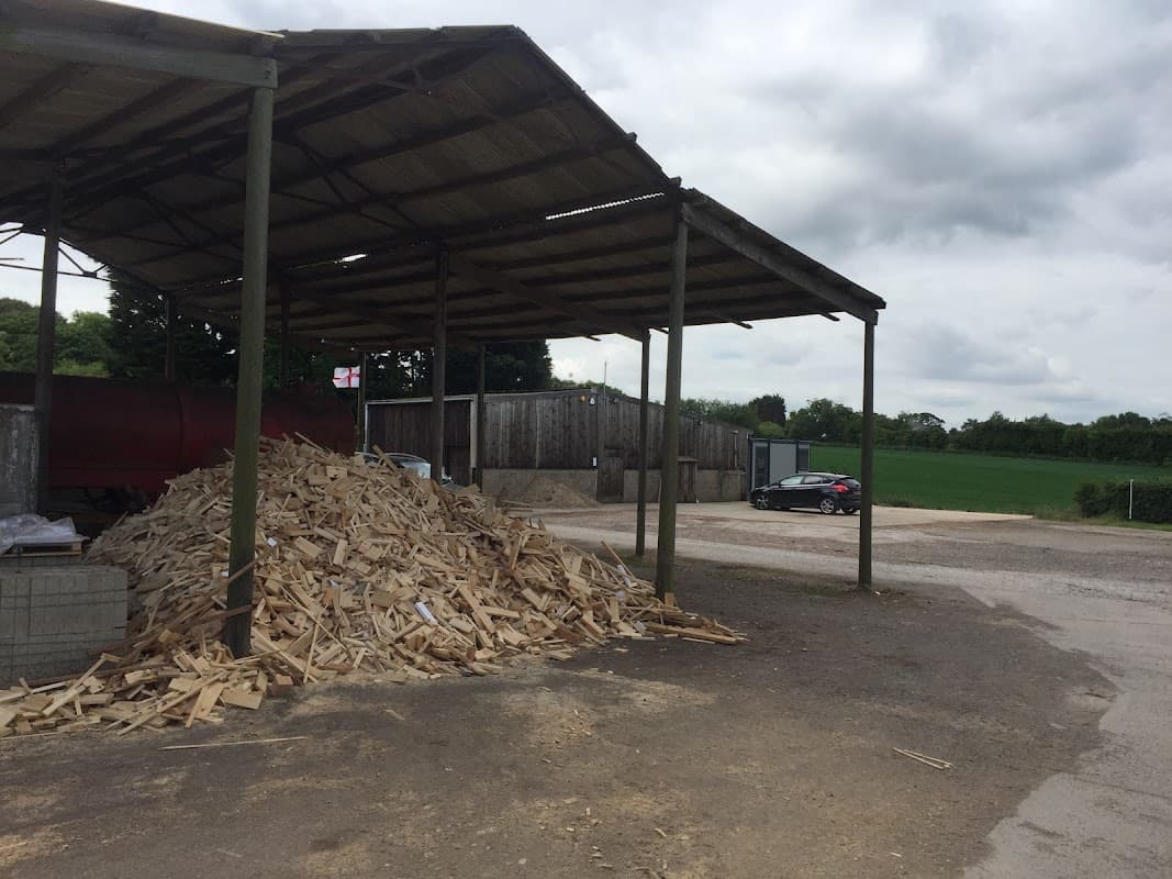 Woodpile under a metal roof, with a gravel area, buildings in the background, and a car parked nearby.