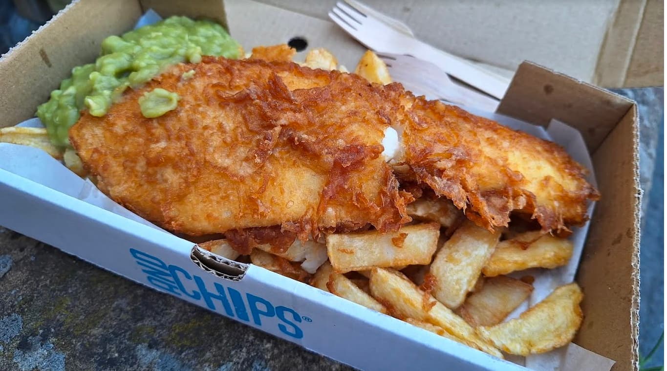 Crispy battered fish served with golden chips and a side of mushy peas in a takeout box.