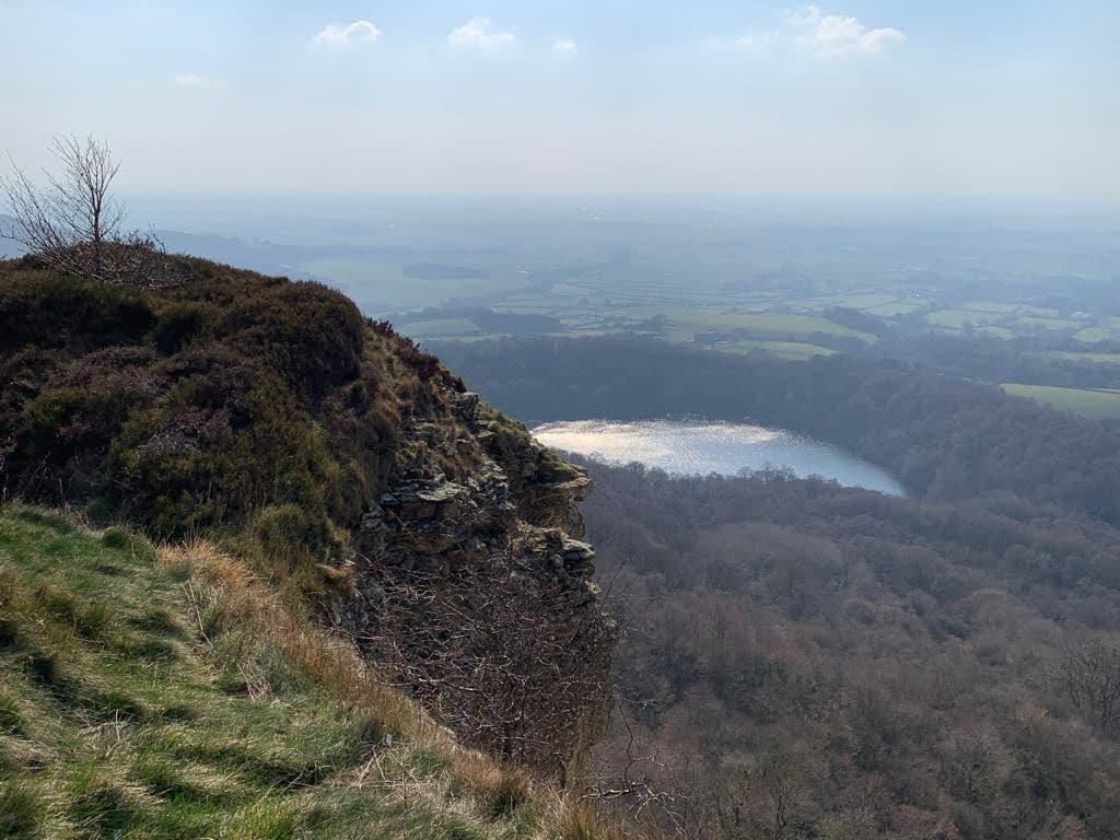 Scenic view from Kilburn Forest Car Park, overlooking a lake surrounded by trees and rolling hills under a hazy sky.