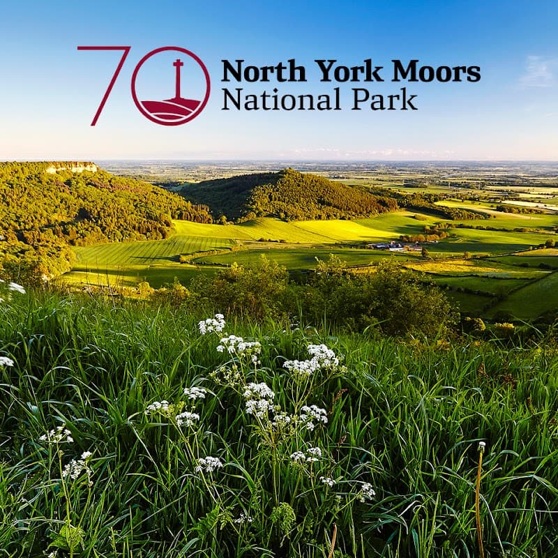 Lush green hills and fields under a clear blue sky at Sutton Bank, with the North York Moors logo in the corner.