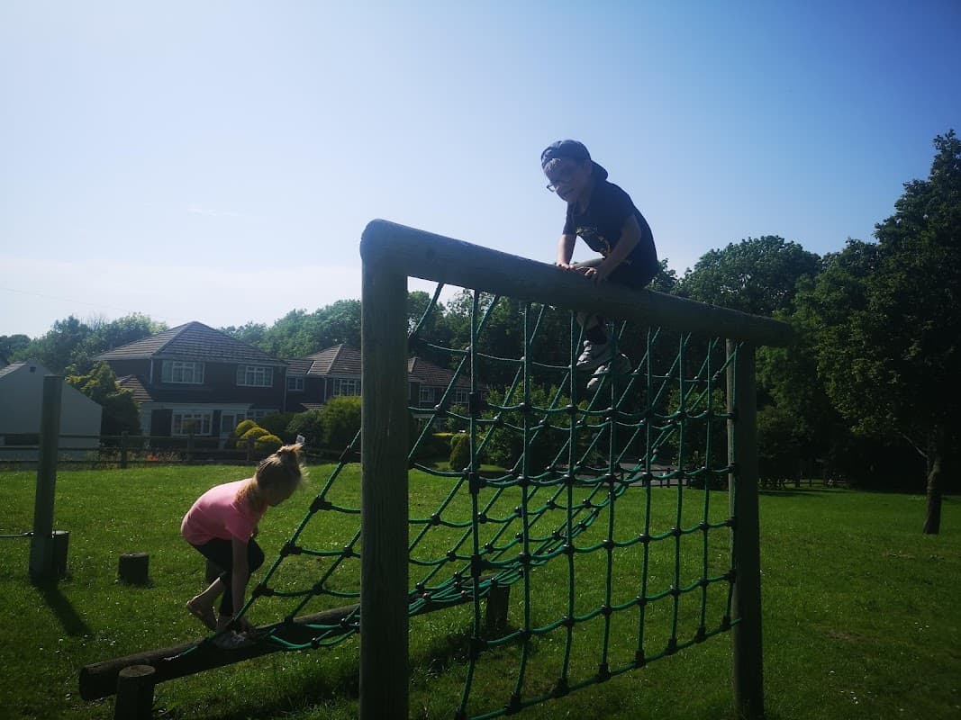 Children playing on a climbing frame in a sunny park, with green grass and houses in the background.