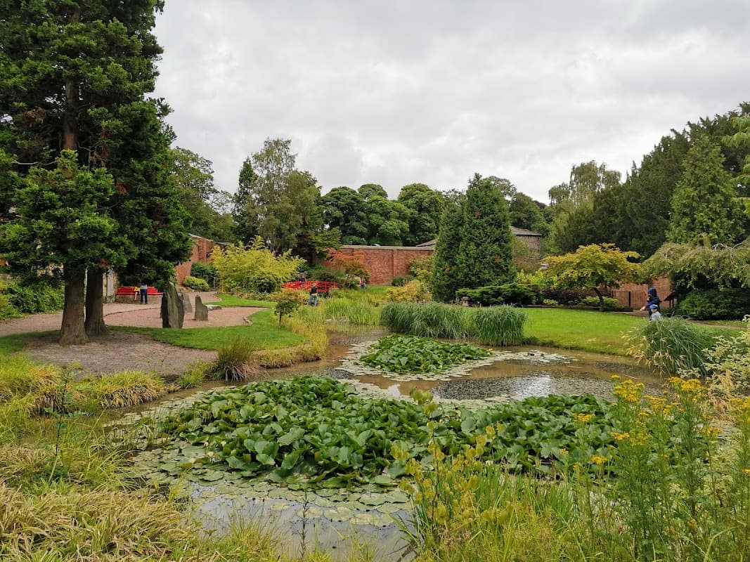 Lush greenery surrounds a tranquil pond with lily pads, set against a cloudy sky in Horsforth Japanese Gardens.