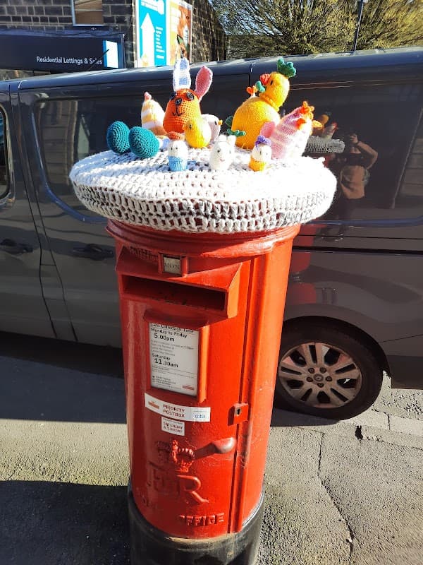 Brightly decorated red post box topped with colorful knitted figures, set against a backdrop of a parked vehicle.