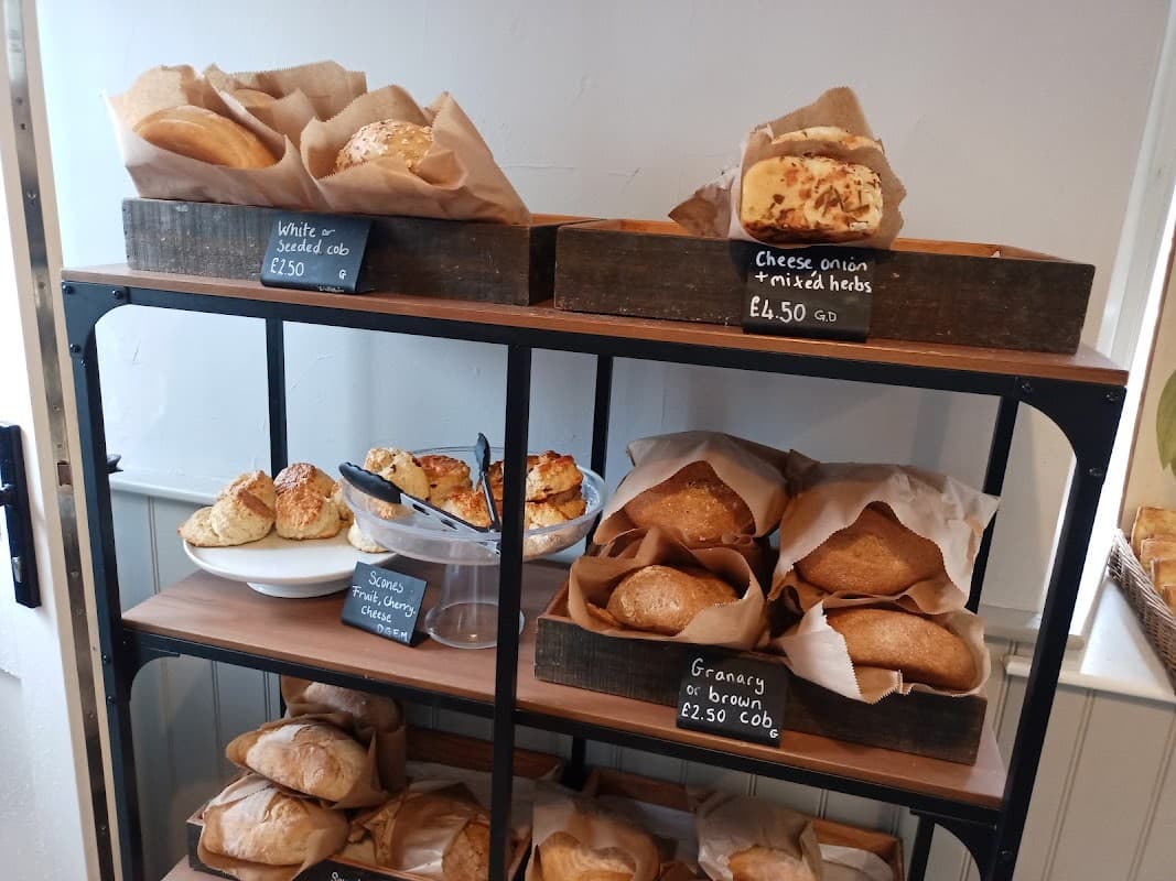 Wooden shelves display various baked goods, including loaves, rolls, and scones, with prices on chalkboard signs.