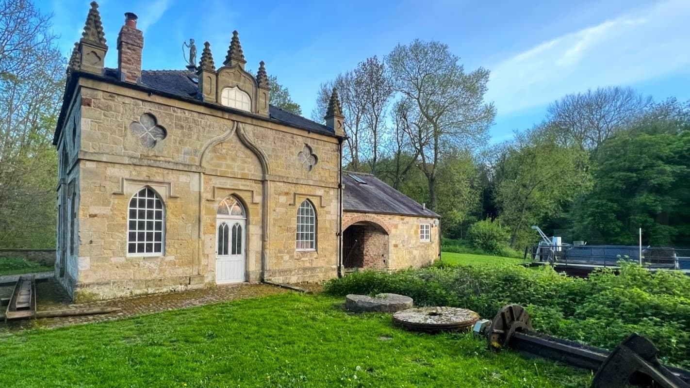 Historic stone building with ornate features, surrounded by greenery and a water wheel, near a tranquil river.