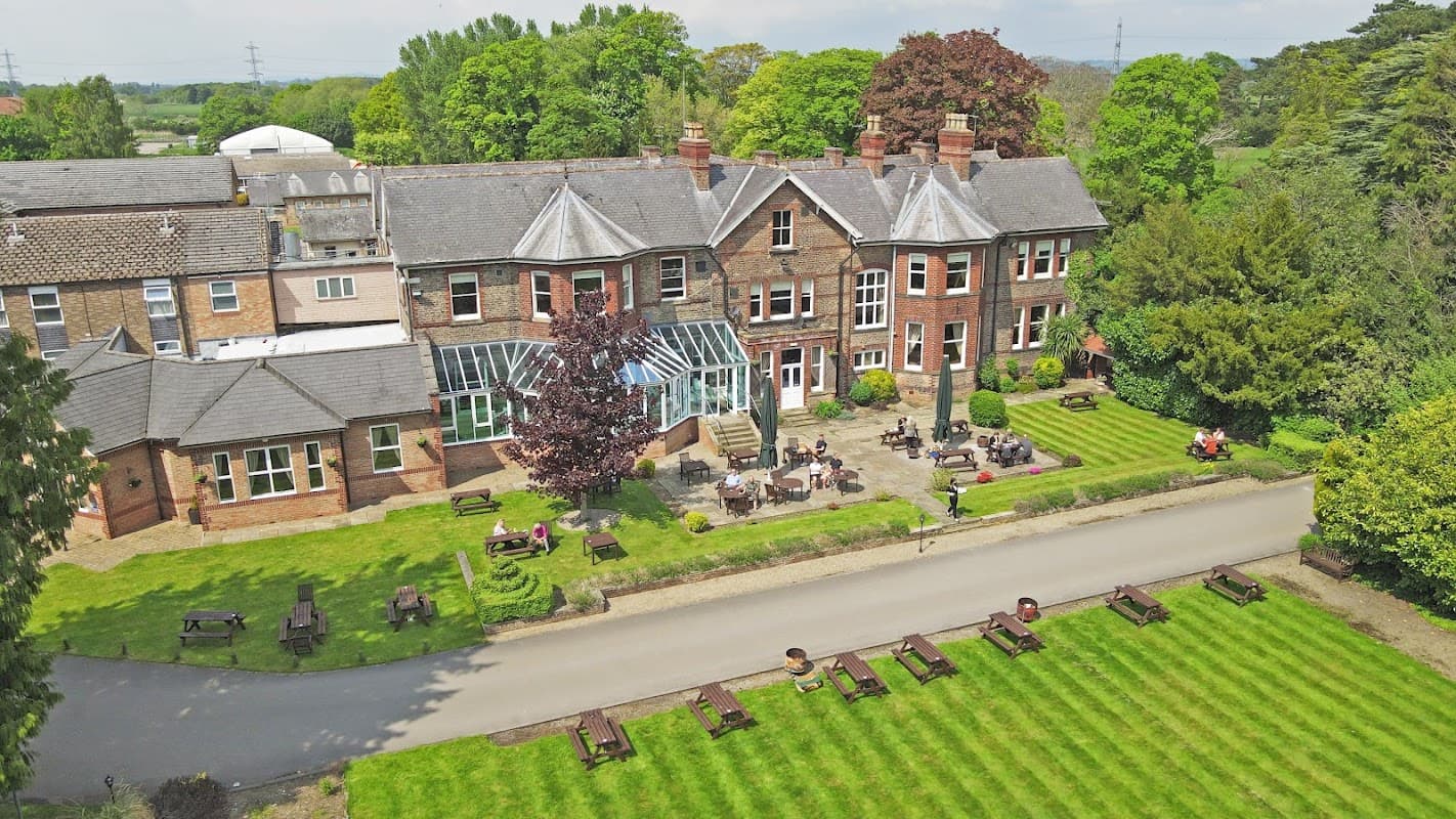 Burn Hall Hotel features a grand brick facade, manicured lawns, outdoor seating, and lush greenery in a serene setting.