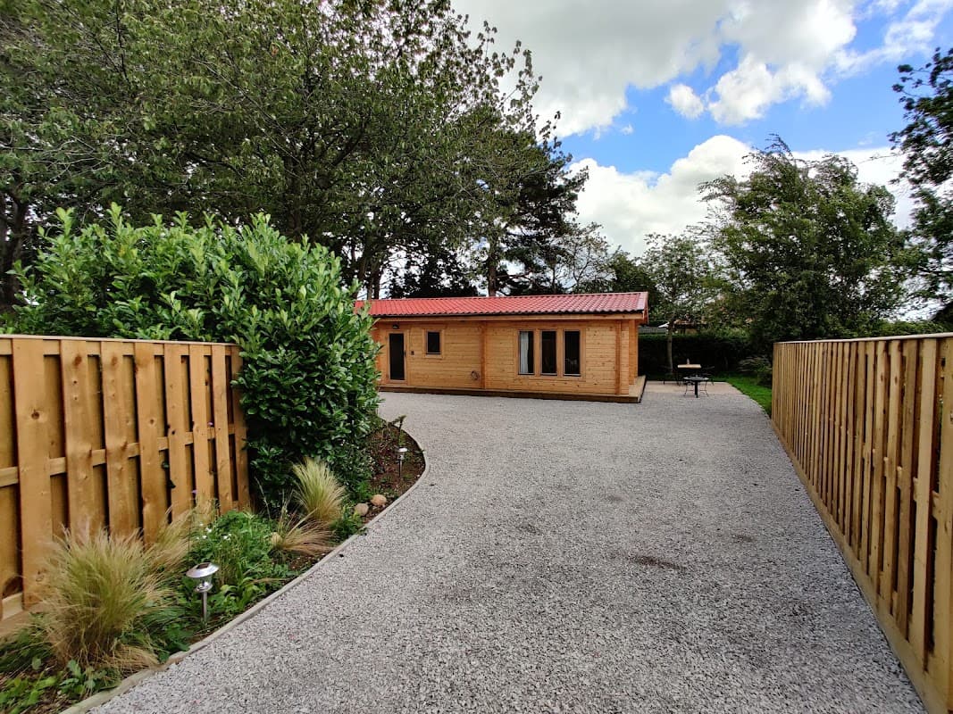 Wooden lodge with red roof surrounded by greenery and gravel pathway, under a partly cloudy sky in Huby, Yorkshire.