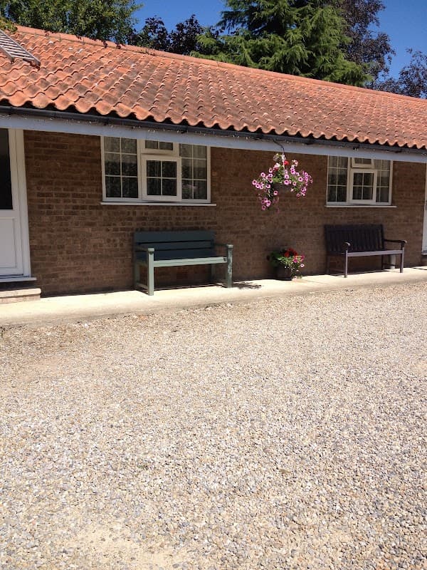 Single-story motel with a tiled roof, gravel pathway, benches, and flower pots in a sunny outdoor setting.