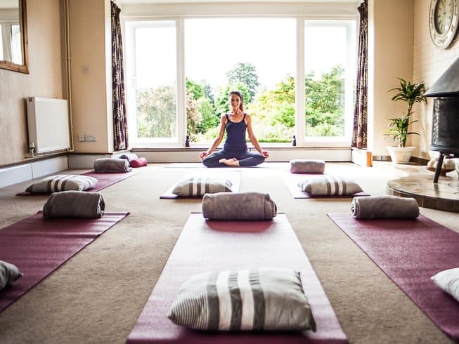 Yoga studio with mats and cushions arranged neatly, a woman meditating in a serene, sunlit space surrounded by greenery.
