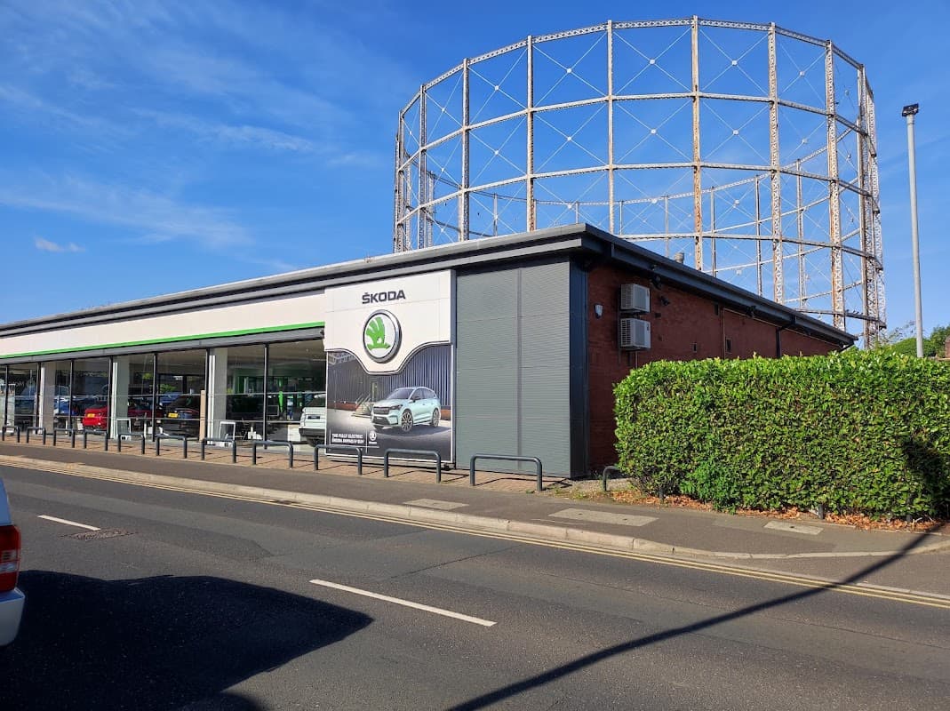 Skoda dealership with a green hedge and a gas holder in the background under a clear blue sky.