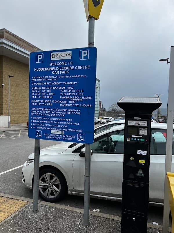 Sign for Huddersfield Leisure Centre Car Park with parking rates and payment machine visible in the foreground.