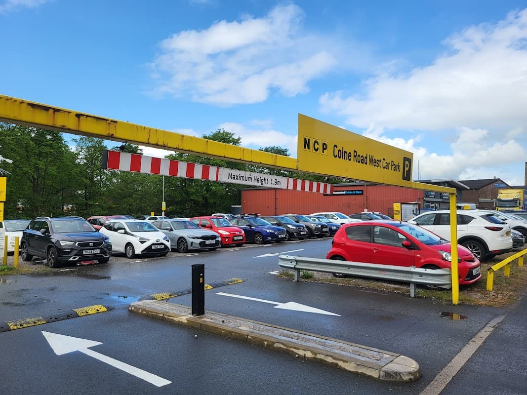 NCP Colne Road car park with parked cars, yellow signage, and trees in the background under a cloudy sky.