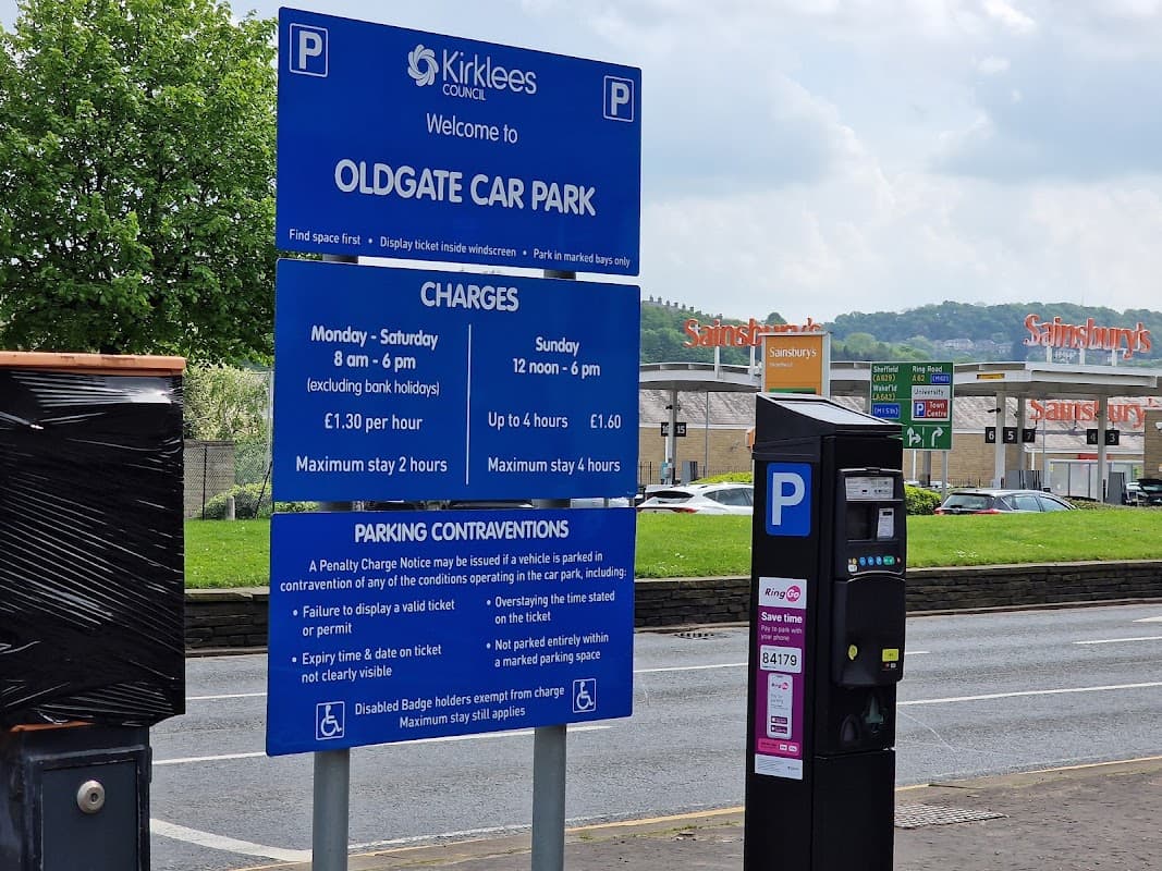Oldgate Car Park sign with parking charges, hours, and payment machine, surrounded by greenery and nearby shops.